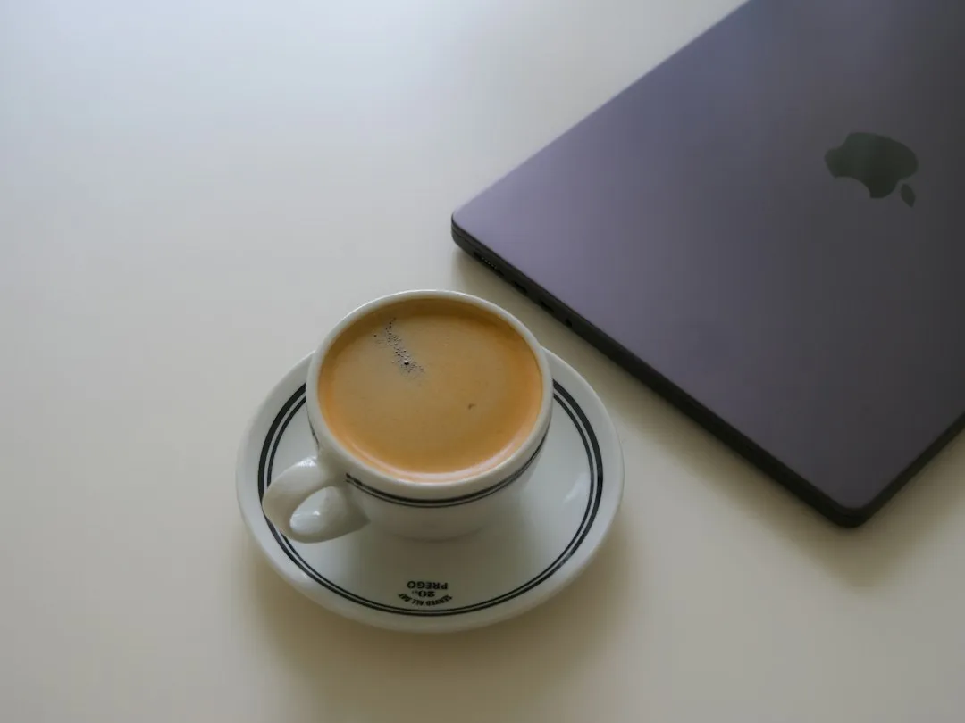 A cup of espresso beside a closed laptop on a white table in morning light.